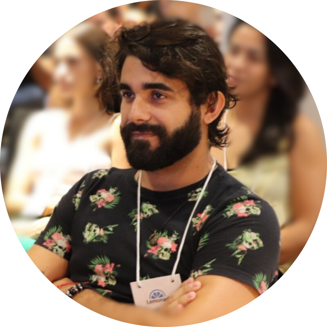 Victor Mourthé, Líder de Tecnologia da CoProdux, sentado e sorrindo, vestindo uma camiseta preta com estampa floral, em um ambiente de conferência com outras pessoas ao fundo.